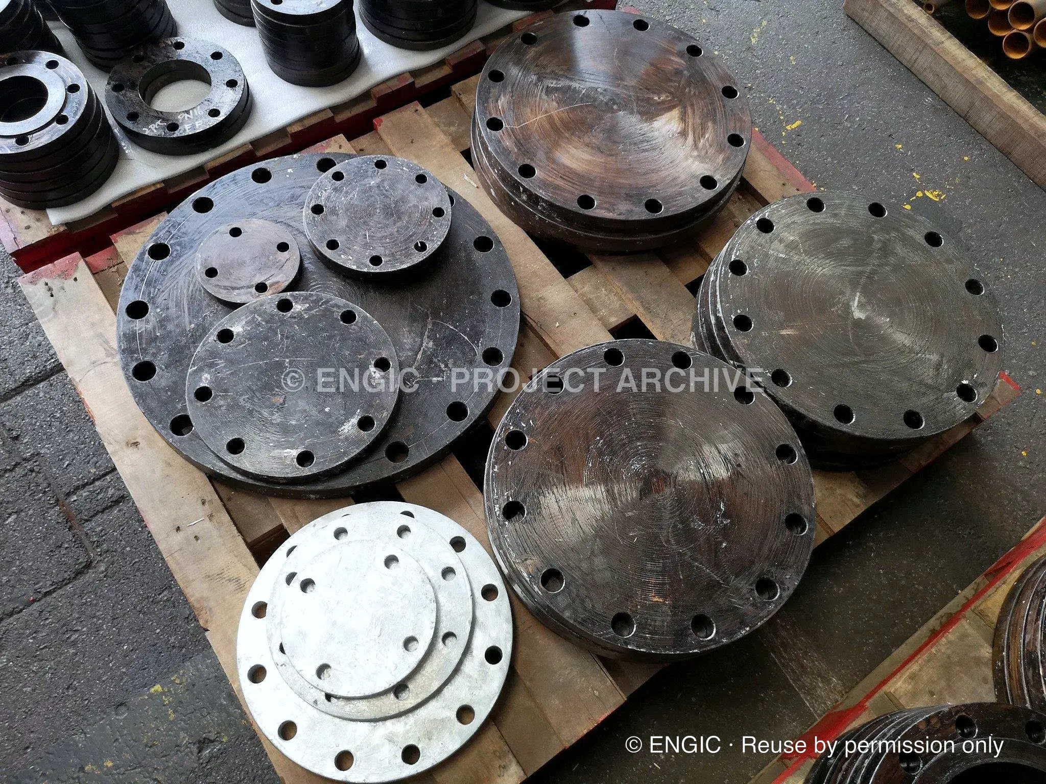 Blind flanges and machined discs stacked on pallets in a yard, with smaller drilled flanges alongside
