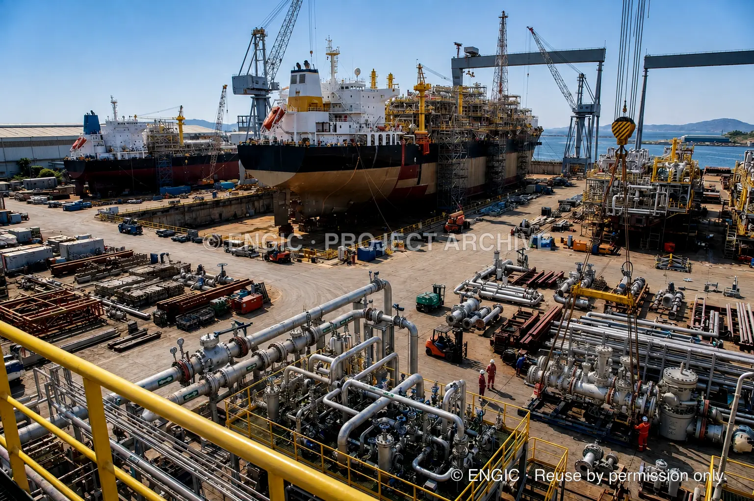 An offshore construction yard with vessels in dry dock, cranes, piping assemblies and workers